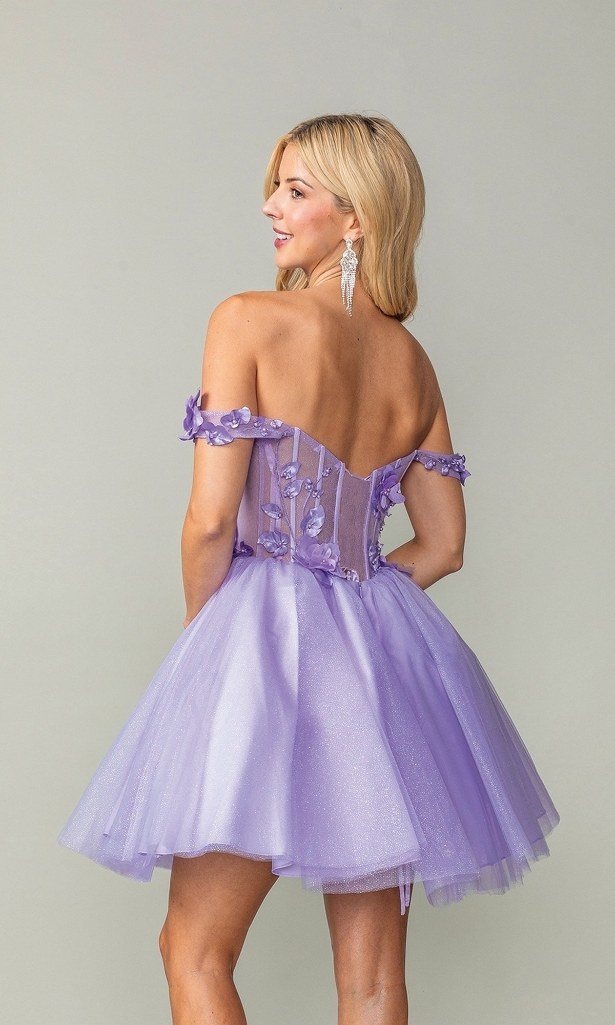 A woman models the Off-Shoulder Short Babydoll Homecoming Dress 3388, a lilac tulle mini dress with floral bodice details, posing with her back to the camera against a plain background.